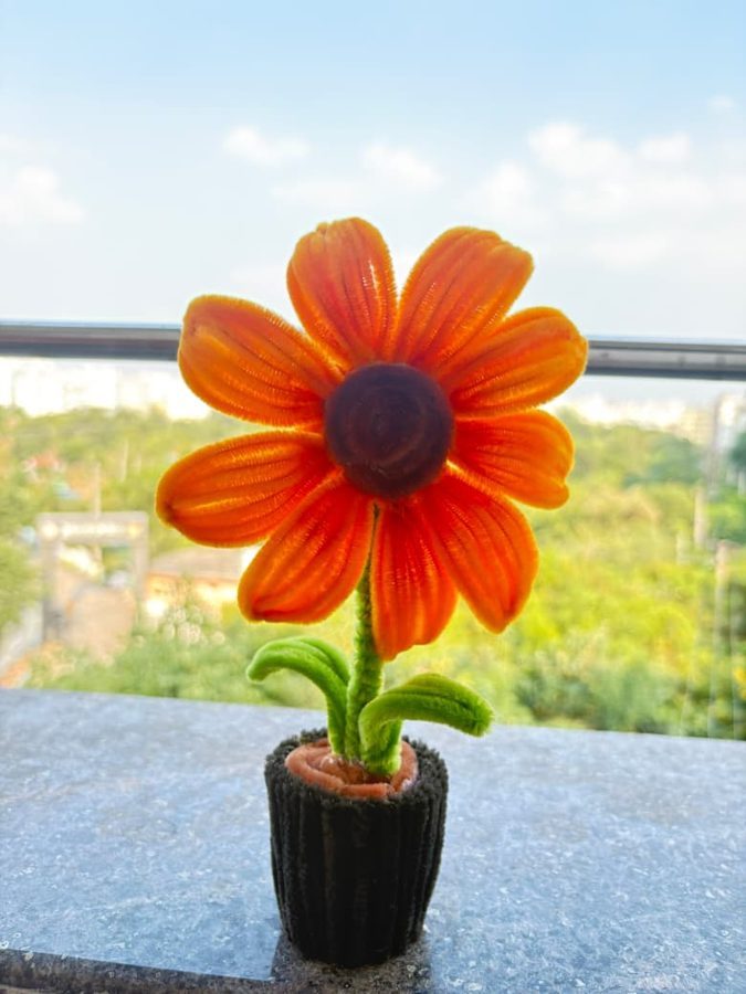 Sunny Petal Pot. Handcrafted orange daisy made from soft chenille wire, with a dark brown center and green leaves. Set in a mini black knitted pot. A cheerful accent for any desk, shelf, or windowsill.